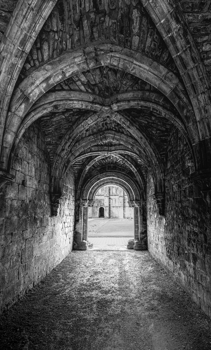 Dark vaulted passage leading towards a bright stone arch